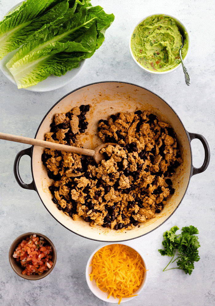 Skillet full of ground turkey and ground beans, surrounded by lettuce, guacamole, salsa, cheese, and cilantro on white table.