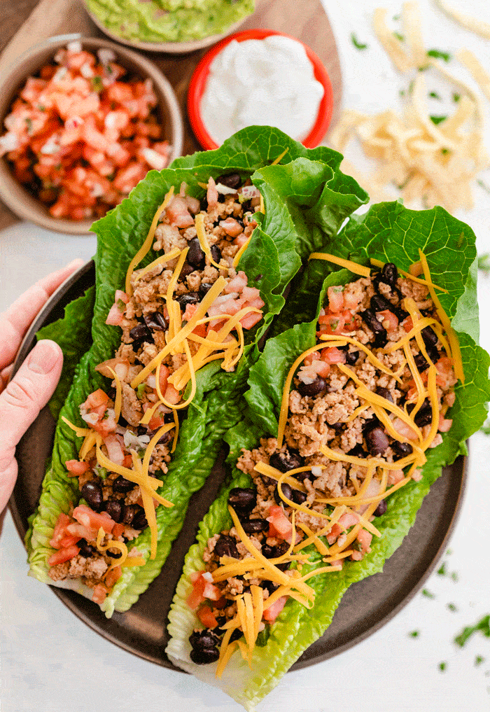 Hand holding a plate of turkey tacos with salsa, sour cream, and guacamole in the background.