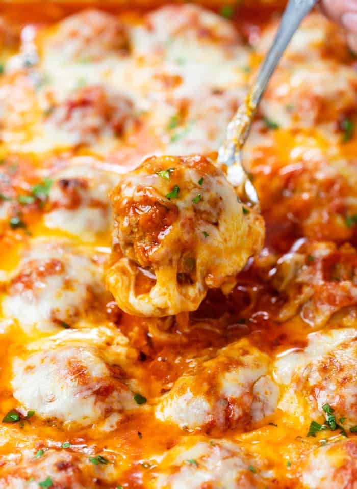 A spoon pulling up a cheesy baked meatball in marinara sauce from a casserole dish.