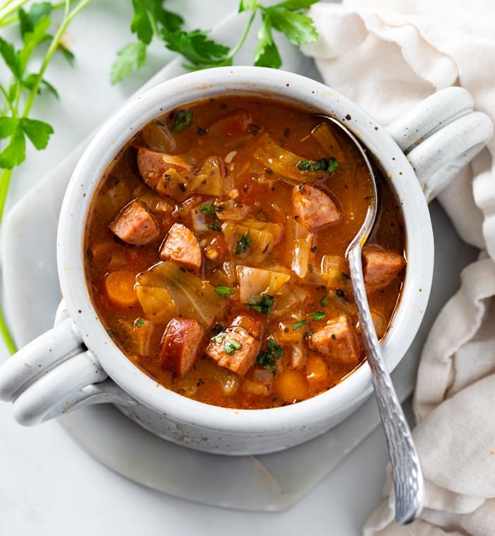 A white crock with Cabbage Soup with a spoon and parsley on the side.