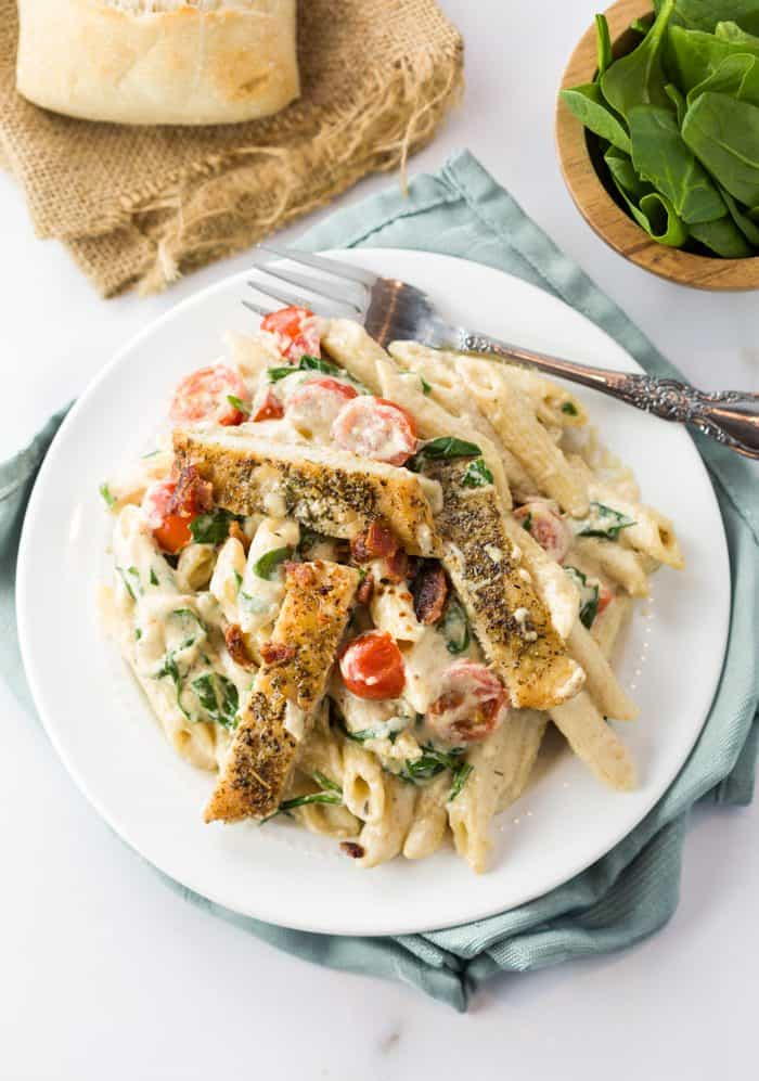 A white plate filled with chicken alfredo pasata on a table next to a roll.