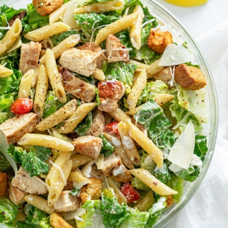 A glass bowl filled with Chicken Caesar Pasta Salad with Lemon wedges on the side.