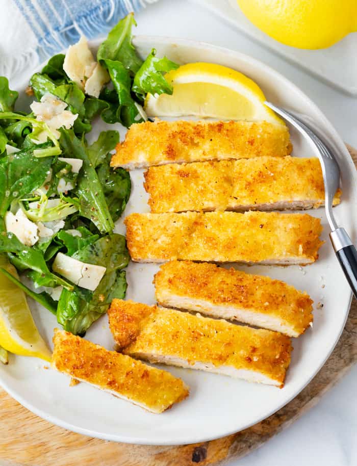 Chicken Milanese cut into slices on a plate with lemon wedges, fresh greens, and Parmesan shavings.