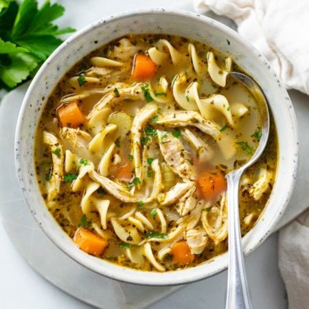 A bowl of chicken noodle soup with a spoon in the bowl and parsley.