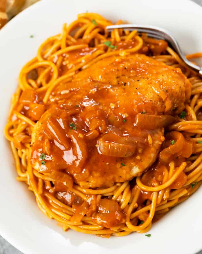 Chicken breast on top of a pile of spaghetti with a red chicken scallopini sauce with sliced onions and parsley.