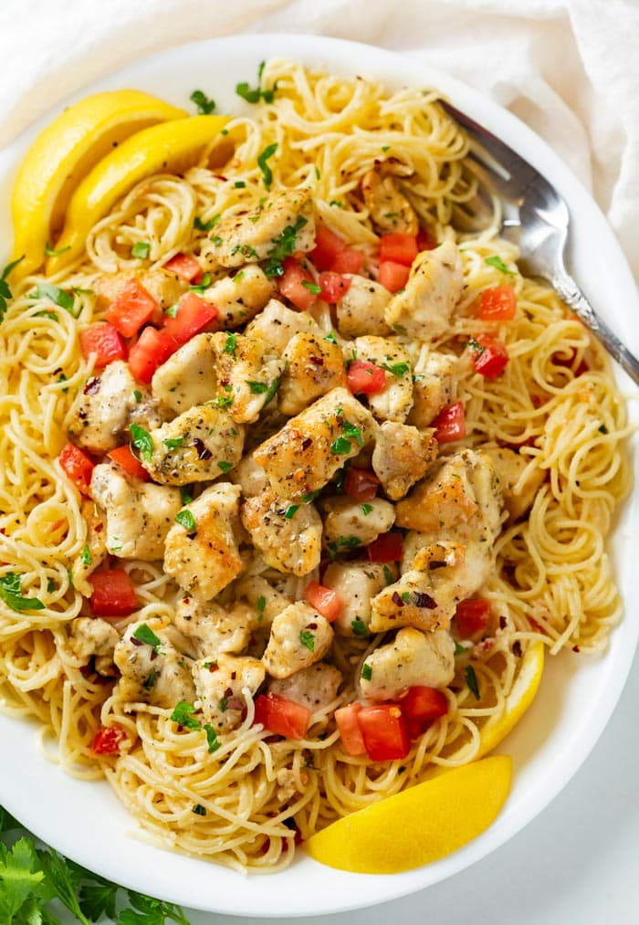 A white platter with Chicken Scampi topped with diced tomatoes, parsley, and slices of lemon.