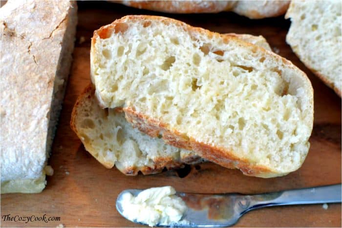 Sliced homemade ciabatta bread on a wooden surface next to a spreader with butter on it.