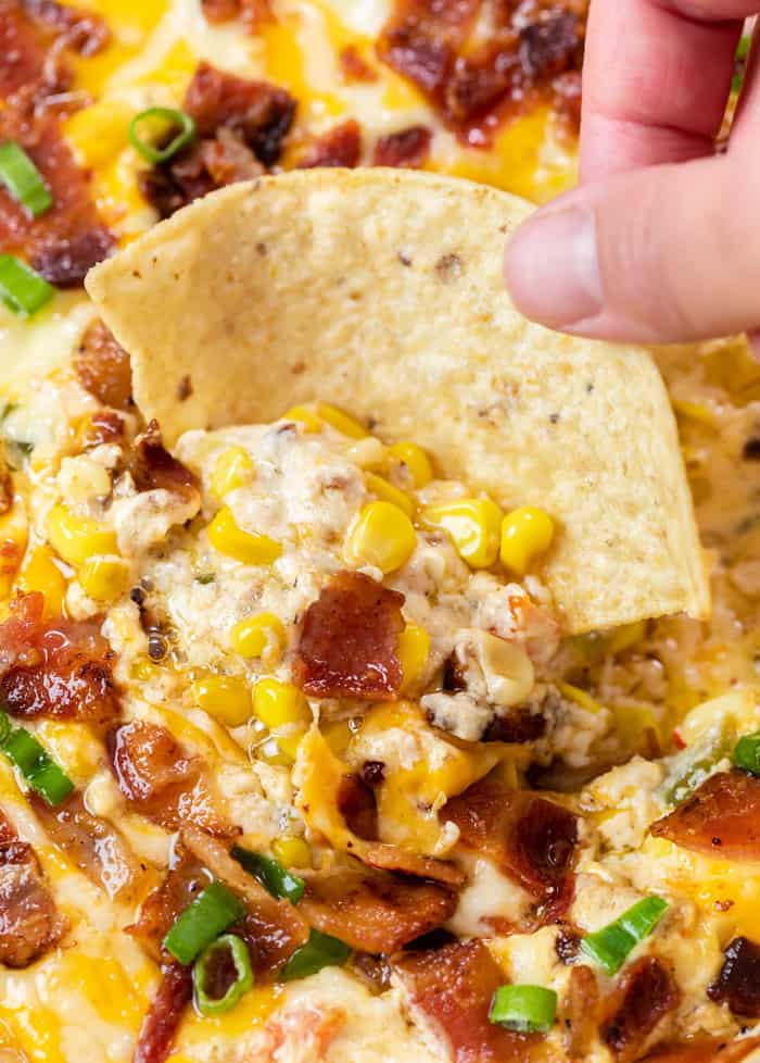 A hand holding a tortilla chip and dipping it into hot corn dip with bacon.