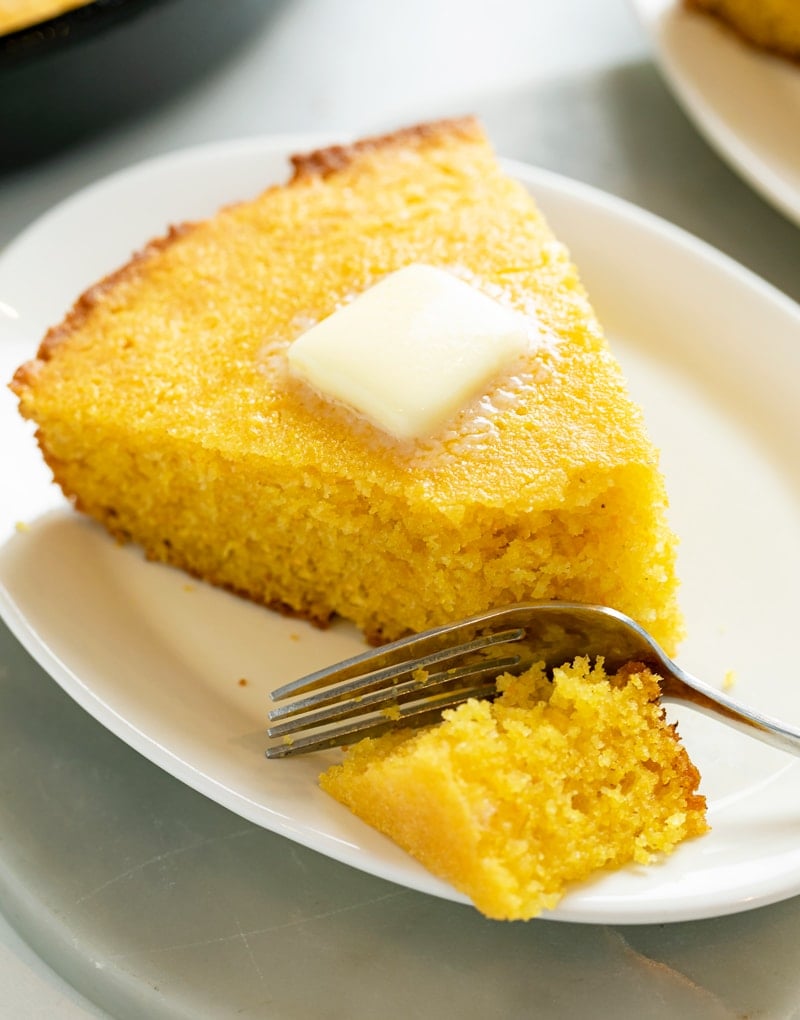 A white plate with a slice of Cornbread with butter on top and a fork.