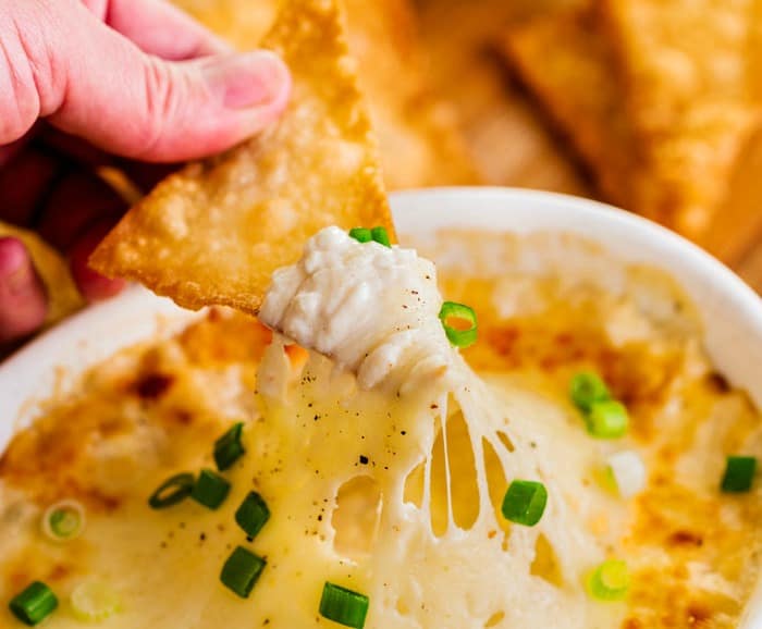 A hand dipping a fried wonton chip into a bowl of crab rangoon dip.