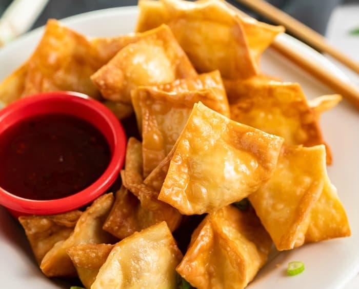 A white plate with a pile of crispy crab rangoon on top with a ramekin of sweet and sour sauce.