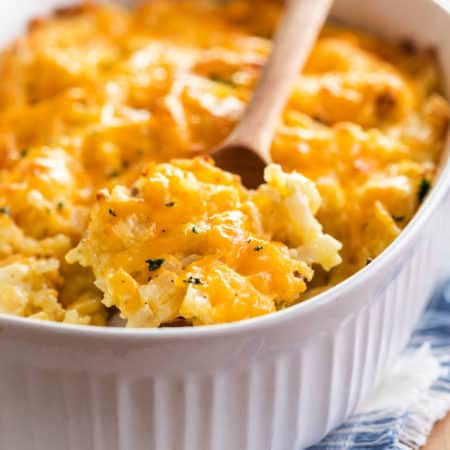 A white casserole dish with a wooden spoon scooping up Cracker Barrel hash brown casserole topped with cheese.