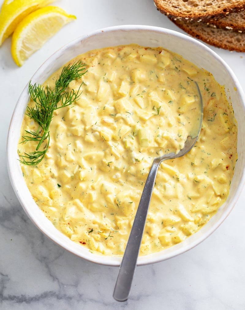 A white bowl filled with egg salad with fresh dill on the side and lemon wedges.