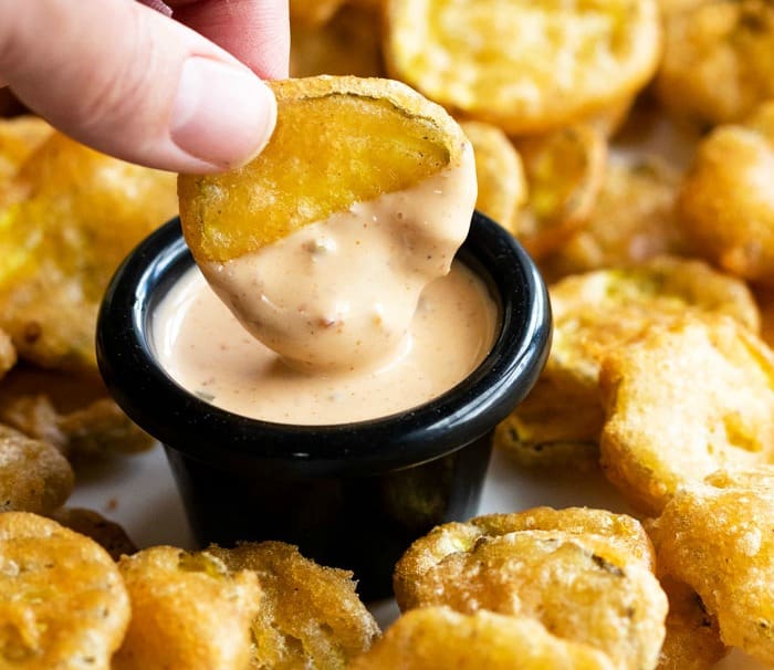 A hand dipping a golden fried pickle into a ramekin of dip.