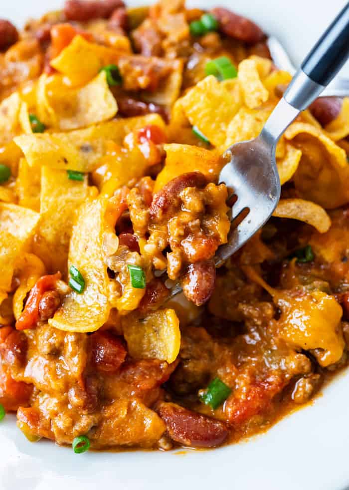 A white plate with Frito Pie and a fork pulling up the cheesy ground beef and beans.