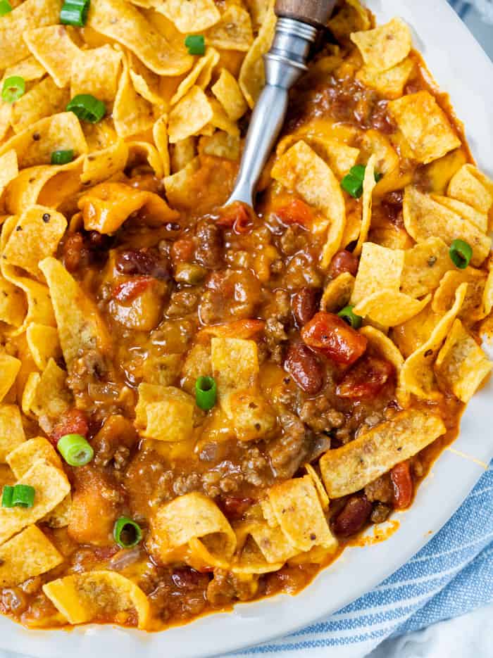 A white pie pan filled with Cheesy Chili topped with Fritos with a spoon in it for Frito Pie.