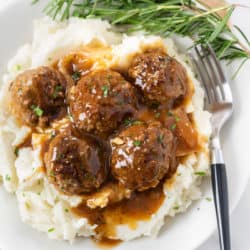 A white plate with a pile of mashed potatoes topped with meatballs and gravy with fresh rosemary on the side.