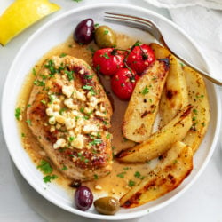 Greek Chicken on a white plate with Feta on top next to potatoes, tomatoes, and olives.