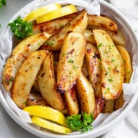 Greek Potatoes in a white bowl with lemon wedges and fresh parsley.