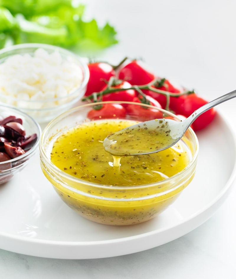 A spoon taking Greek Salad Dressing from a glass cup on a white plate.