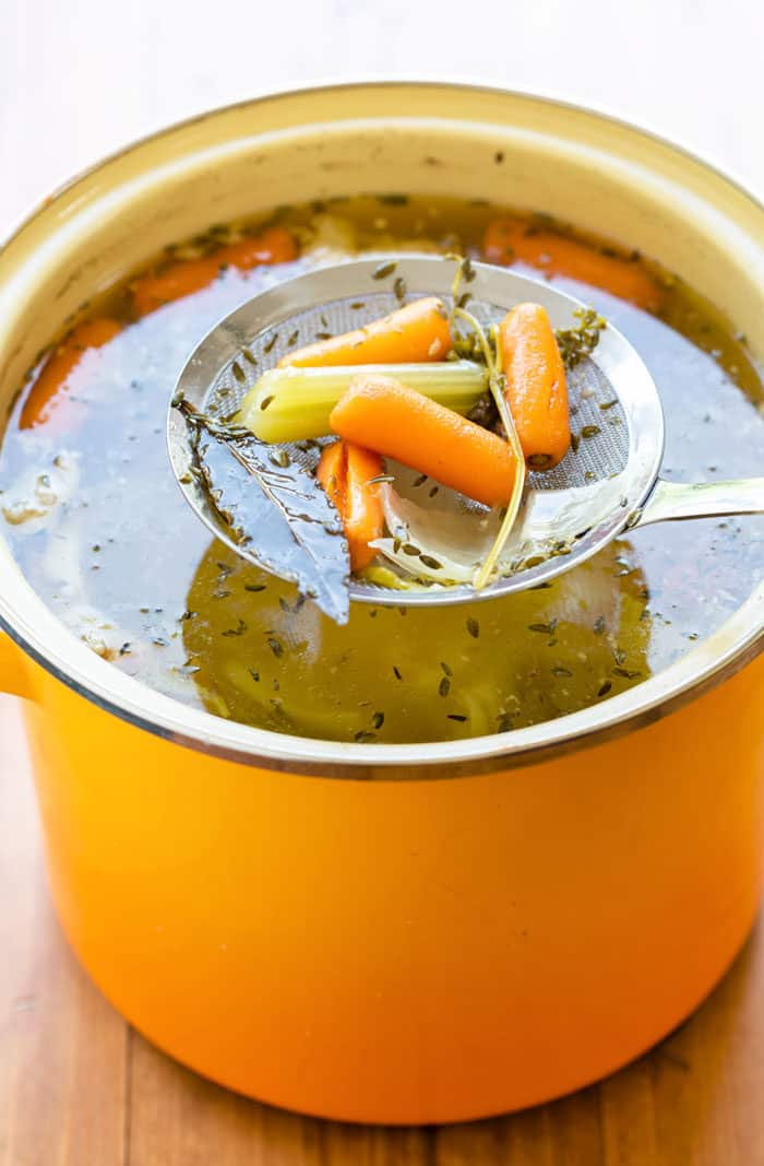 A skimmer spoon being dipped into a bowl of chicken stock to remove the vegetables.