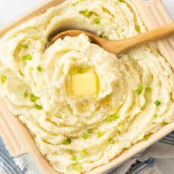 Overhead view of a casserole dish and a wooden spoon scooping up creamy homemade mashed potatoes topped with butter.