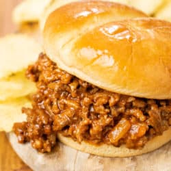 A hamburger bun filled with homemade sloppy joes on a wooden plate with chips in the background.