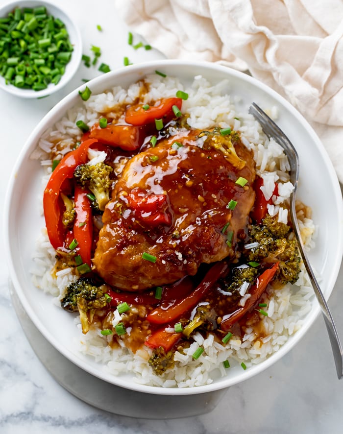 A white plate Honey Garlic Chicken on top of rice with Broccoli and Red Peppers.