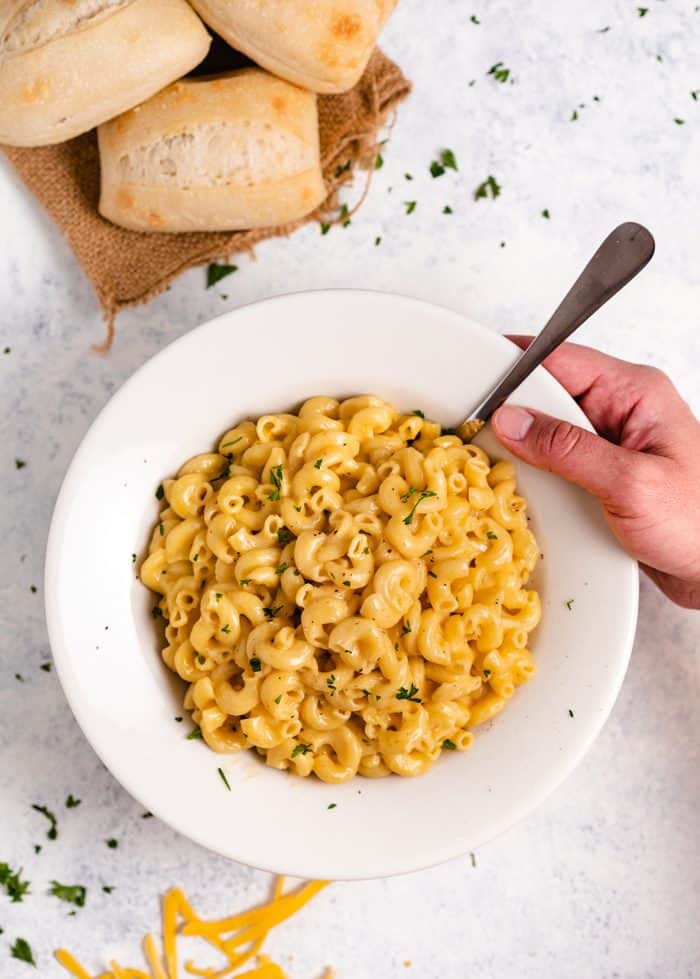 A hand reaching for a white bowl filled with macaroni and cheese on a light white surface with rolls in the background.