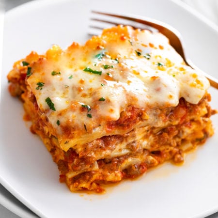 A white plate with Lasagna on it with a fork in the background.