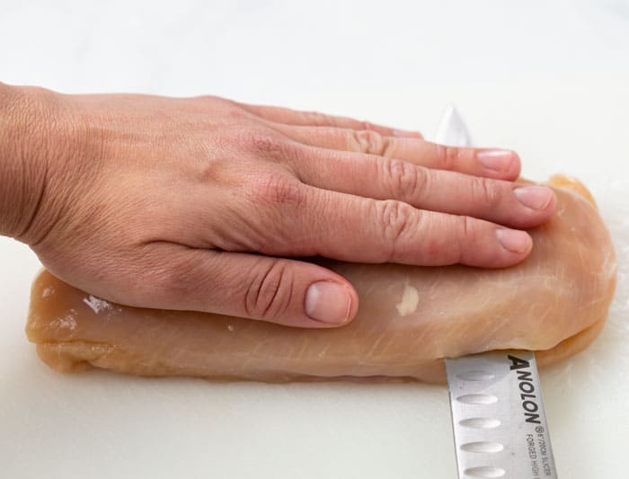 A hand over a chicken breast with a knife cutting it in half.