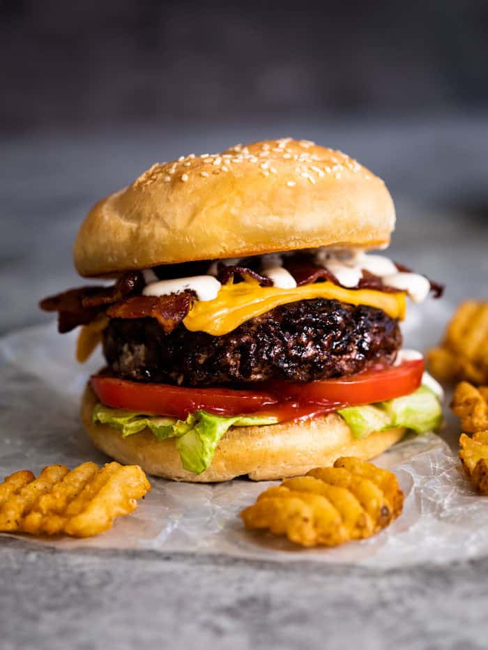 A bacon cheeseburger in between a bun with lettuce and tomatoes, drizzled with ranch dressing. Waffle fries in the front.