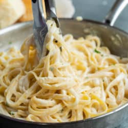 Kitchen tongs pulling Fettuccine Alfredo up from a pan.