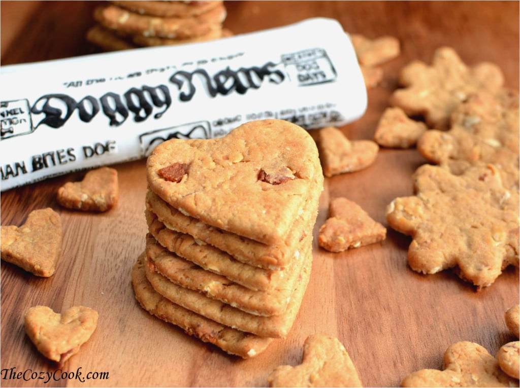 A stack of heart shaped dog greats on a wooden surface surrounded by smaller dog treats with a toy newspaper in background.