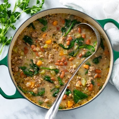 A green Dutch oven filled with Pinto Bean Soup with a soup ladle in it and fresh parsley on the side.