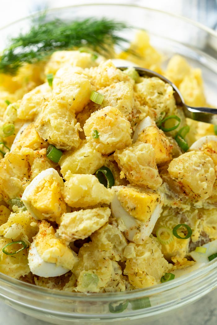 A glass bowl with potato salad with hardboiled eggs and fresh dill in the back.