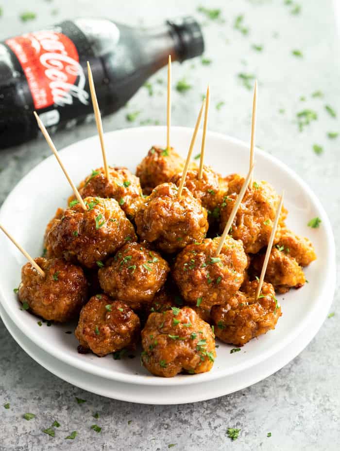 A white plate filled with brown glossy spicy sausage bites with a coca cola bottle in the background. 