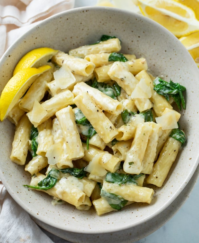 A white bowl filled with Creamy Spinach Pasta with ziti noodles and lemon wedges on the side.