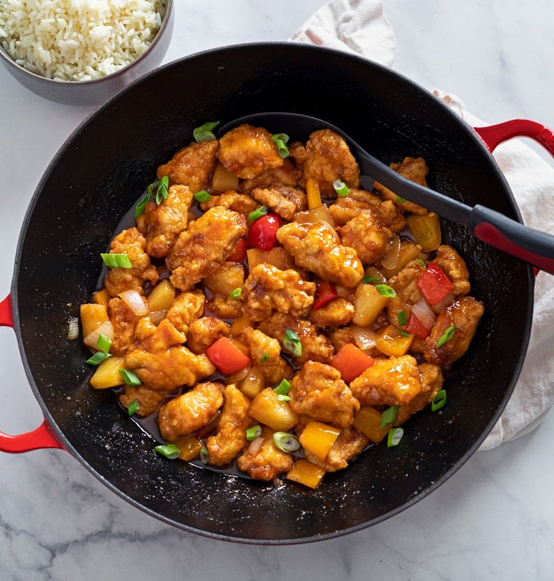 A wok filled with Sweet and Sour Chicken with green onions and white rice on the side.