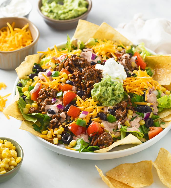 A white plate filled with Taco Salad topped with ground beef, avocado, sour cream, tortilla chips, and vegetables.