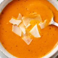 A bowl of Tomato Soup with a spoon in it and shaved Parmesan Cheese on top.