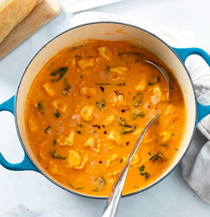 A Dutch oven filled with creamy Tomato Tortellini Spinach soup with spinach and a ladle on the side.