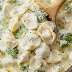 A wood spoon scooping up Tortellini Alfredo with broccoli from a skillet.