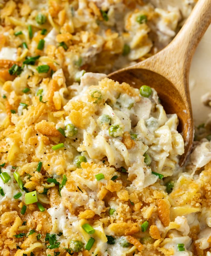 A wooden spoon scooping up creamy Tuna Noodle Casserole with egg noodles and peas.