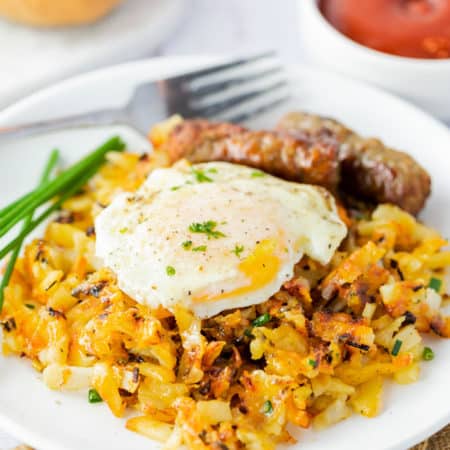 A white plate with waffle iron hash browns topped with a fried egg with sausage links in the background.