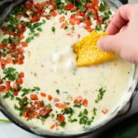 A hand dipping a tortilla chip into White Queso Dip garnished with Pico De Gallo and cilantro.